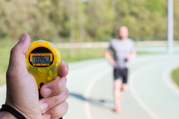 Hand hält eine gelbe Stoppuhr mit 00:08:66, während ein Jogger auf einer Laufbahn im Hintergrund läuft.