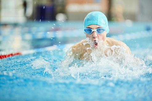 Junger Schwimmer im Wasser, trägt eine blaue Schwimmkappe und Brille, spritzt Wasser beim Wettkampfschwimmen.
