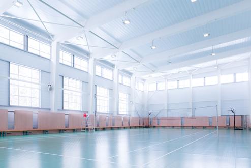 Leere Sporthalle mit hellem Boden, Fensterfront und Volleyballnetzen an der Seite. Eine Leiter steht neben dem Netz.